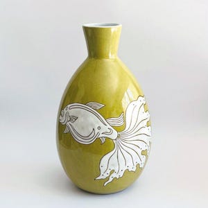 Puede incluir: Un jarrón de cerámica de color chartreuse con un diseño de pez blanco. El jarrón tiene un cuerpo redondeado y un cuello estrecho. El pez está representado con aletas detalladas y una cola fluida, agregando un toque decorativo.