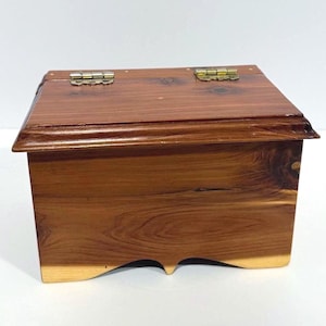 Può includere: Una piccola scatola rettangolare in legno con coperchio incernierato. La scatola è realizzata in legno marrone rossiccio con venature visibili e cerniere in ottone. Il bordo inferiore presenta un design decorativo smerlato. La scatola è probabilmente utilizzata per riporre oggetti o per l'esposizione.