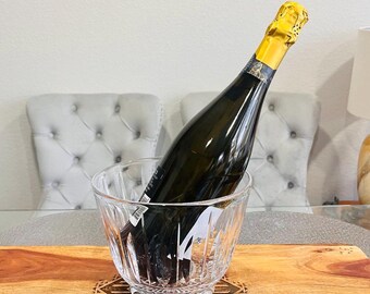 May include: A clear glass champagne bucket with a bottle of champagne inside. The bucket is sitting on a wooden serving tray with a decorative design.