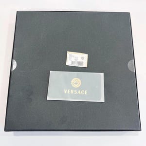 May include: A black square box with a small white tag and a gold and white Versace card. The Versace card has the brand's logo and the word "VERSACE" in gold lettering. The box is closed.