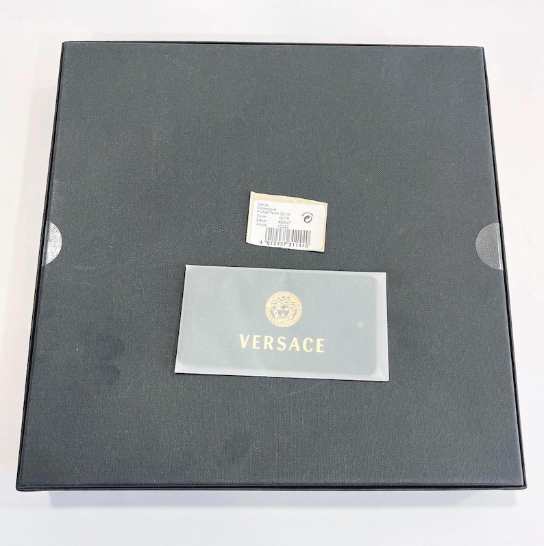 May include: A black square box with a small white tag and a gold and white Versace card. The Versace card has the brand's logo and the word "VERSACE" in gold lettering. The box is closed.