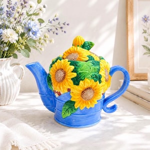 May include: A ceramic teapot shaped like a blue watering can, featuring three-dimensional yellow sunflowers with brown centers and green leaves. The teapot has a curved blue handle, a matching blue spout, and a lid topped with a small yellow bud.