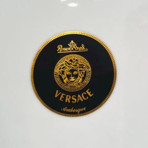 May include: A round black and gold Versace logo with the text "Versace Arabesque" and the Rosenthal logo. The logo features a gold Medusa head in the center.