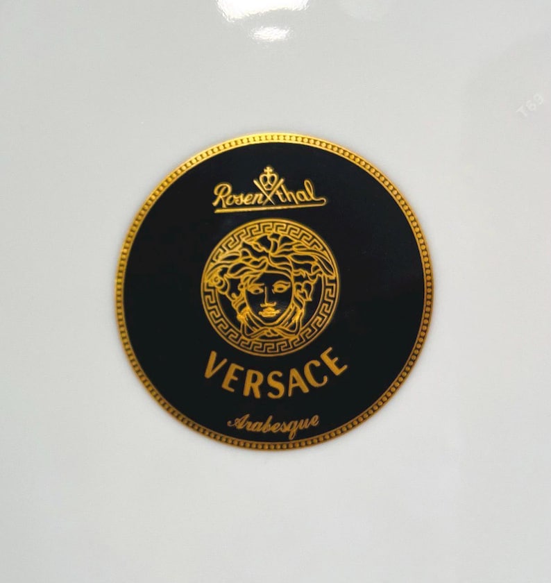 May include: A round black and gold Versace logo with the text "Versace Arabesque" and the Rosenthal logo. The logo features a gold Medusa head in the center.