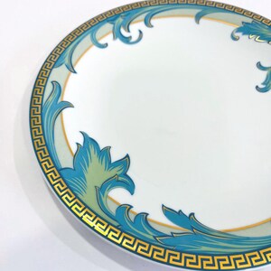 May include: White ceramic plate with a decorative border featuring a gold Greek key design and teal and blue floral accents. The plate has a gold rim and a white center.