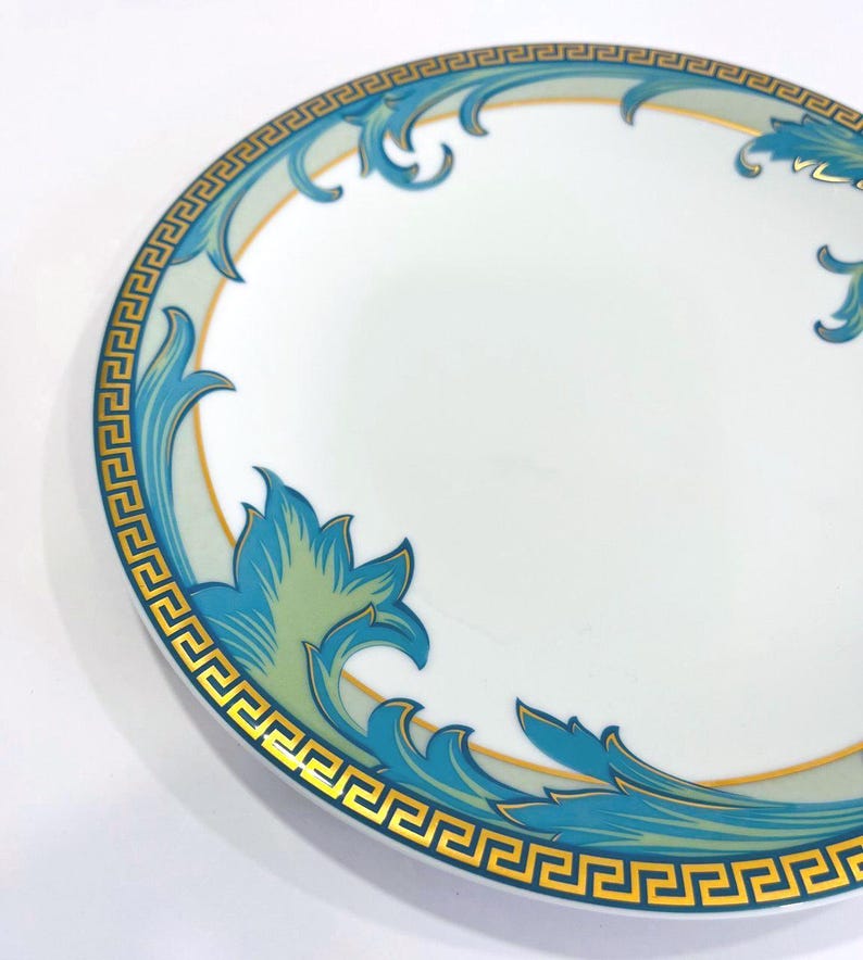 May include: White ceramic plate with a decorative border featuring a gold Greek key design and teal and blue floral accents. The plate has a gold rim and a white center.