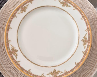May include: A white ceramic plate with a gold and pink floral border. The plate is on a woven placemat.