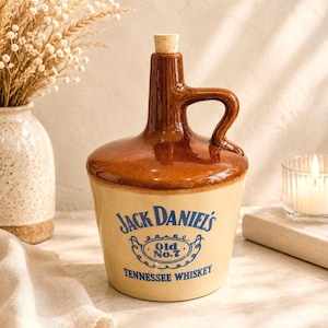 Könnte beinhalten: Ein Keramikkrug mit braun glasierter Oberseite und cremefarbenem Unterteil, der den blauen Schriftzug Jack Daniel's Old No. 7 Tennessee Whiskey trägt. Der Krug hat einen seitlichen Henkel und ist oben mit einem kleinen, hellen Korken verschlossen.