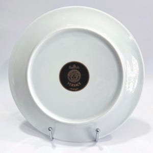 May include: A white porcelain plate with a black and gold Versace logo on the bottom. The plate is round and has a simple, elegant design. The plate is on a silver metal stand.