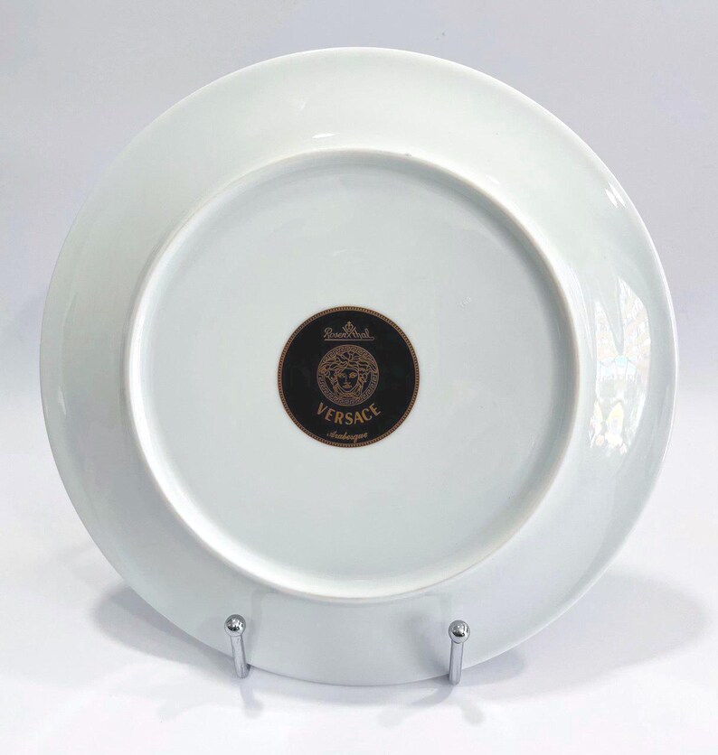 May include: A white porcelain plate with a black and gold Versace logo on the bottom. The plate is round and has a simple, elegant design. The plate is on a silver metal stand.