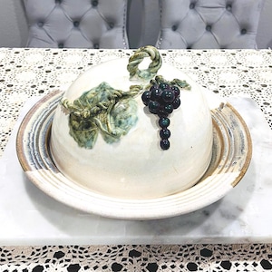 Puede incluir: Una mantequera de cerámica con tapa, con un diseño de uvas y hojas. El plato blanquecino tiene un borde marrón y se encuentra sobre una superficie de mármol. La tapa tiene un asa retorcida y un racimo de uvas.