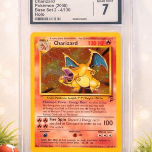 May include: A graded Charizard Pokémon card from the Base Set 2 - 4/130 Holo set, graded Near Mint 7 by CGC. The card features a vibrant illustration of Charizard, with text detailing its abilities and stats. The card is encased in a protective holder.