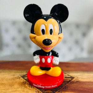 K&ouml;nnte beinhalten: Eine gelbe und schwarze Mickey Mouse Bobblehead mit rotem Sockel. Der Sockel tr&auml;gt den Schriftzug "Walt Disney World Resort".