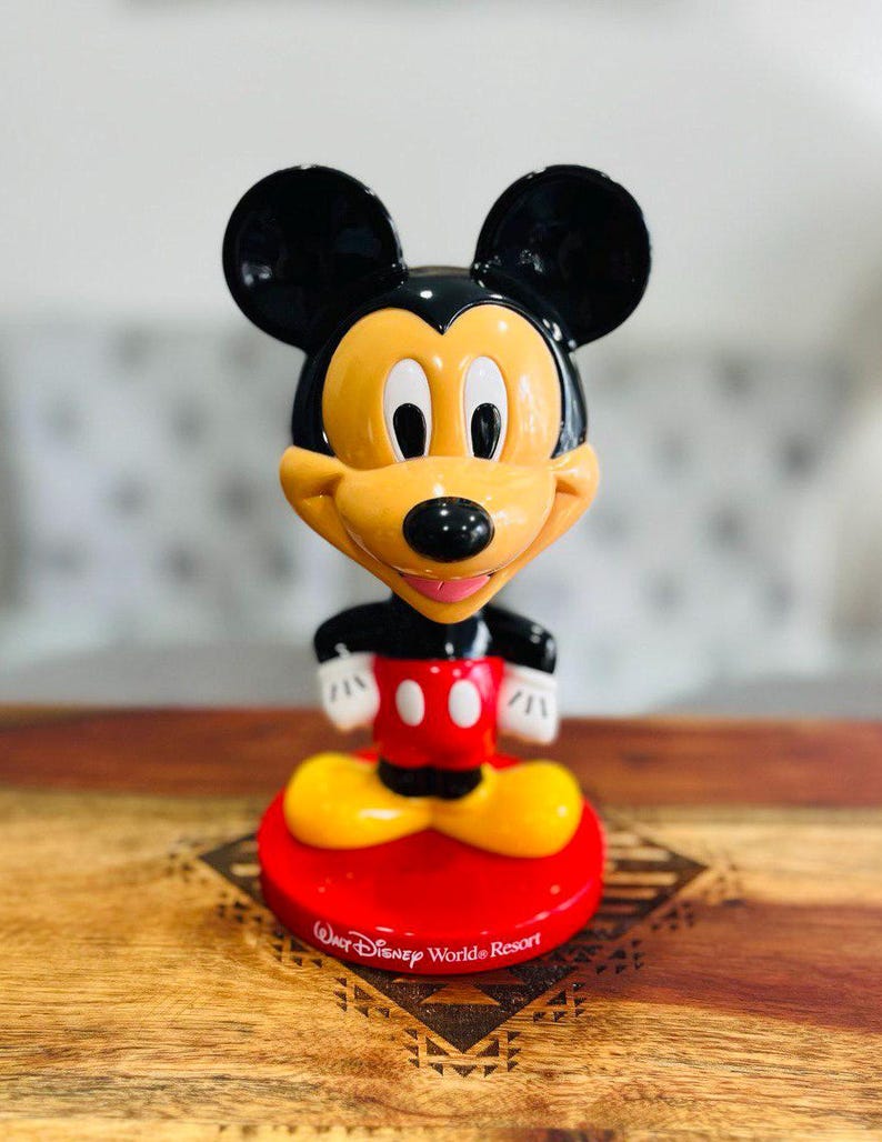 K&ouml;nnte beinhalten: Eine gelbe und schwarze Mickey Mouse Bobblehead mit rotem Sockel. Der Sockel tr&auml;gt den Schriftzug "Walt Disney World Resort".