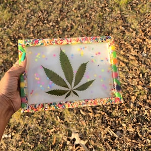 May include: A rectangular clear tray with a colorful sprinkle border. Inside, a large green cannabis leaf is displayed with colorful confetti. The tray is held by a hand.