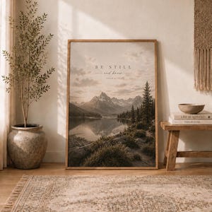 Op de afbeelding: Een rechthoekige ingelijste wandkunstprint met een landschap van een kalm meer dat besneeuwde bergen en dennenbomen onder een bewolkte lucht weerspiegelt. De tekst BE STILL and know PSALM 46:10 staat in het midden bovenaan.