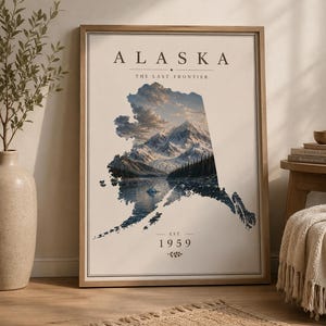 May include: A framed wall art print featuring the outline of Alaska filled with a landscape of snow-capped mountains, a dark forest, and a reflective lake. The text reads ALASKA, THE LAST FRONTIER, and EST. 1959, centred on a cream-coloured background.