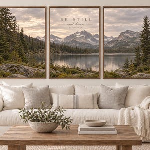 Puede incluir: Un conjunto de tres cuadros de pared que muestra un paisaje panorámico de un lago de montaña con pinos y un cielo al atardecer. El panel central muestra el texto BE STILL and know PSALM 46:10 en una fuente oscura sobre el paisaje.