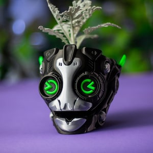 May include: A black and silver robotic-style planter featuring two prominent circular eyes with glowing green accents. The container has a mechanical, segmented design and holds a small, light-colored plant with jagged, textured leaves.