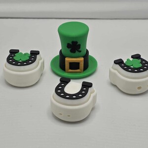May include: Four white ceramic charms with green shamrock and black horseshoe designs. One charm is a green leprechaun hat with a black band and gold buckle.