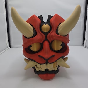 May include: A red and black Oni mask with tan horns and teeth. The mask has gold-colored eye and forehead details. The mask is a 3D-printed item, likely for cosplay or decoration.