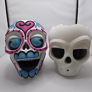 May include: Two decorative skull figurines. One is white with black eye sockets and a small nose opening. The other is white with pink, blue, and black accents, featuring a heart design on the forehead and blue eye details.