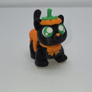 May include: A black cat figurine with large green eyes, wearing an orange pumpkin costume. The cat has a pumpkin hat and a pumpkin-shaped harness. The figurine is a Halloween-themed decoration.