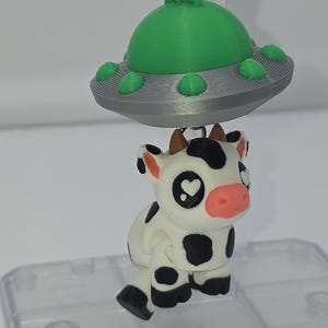 May include: A white and black cow with pink nose and heart-shaped eyes is hanging from a silver and green UFO charm.