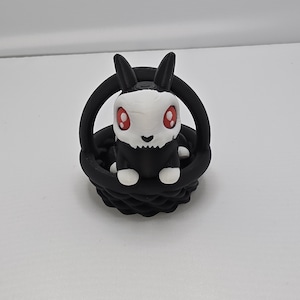May include: Black and white bunny figurine with red eyes sitting in a black basket. The bunny has a skull-like face.