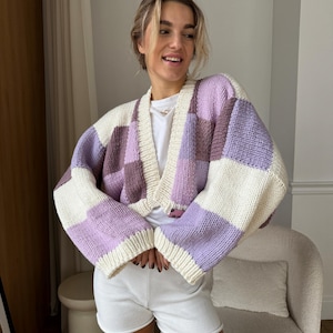 May include: A knitted cardigan with a patchwork design. The sweater features large blocks of lavender, cream, and mauve. The cardigan has long sleeves and a V-neck. The model is wearing a white t-shirt and white shorts.