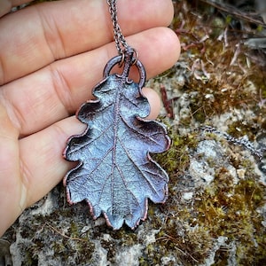 Può includere: Un ciondolo a forma di foglia di quercia con una finitura metallica scura e bordi color rame. La foglia presenta una superficie dettagliata e testurizzata con venature visibili ed è appesa a una sottile catena a maglie in metallo scuro.