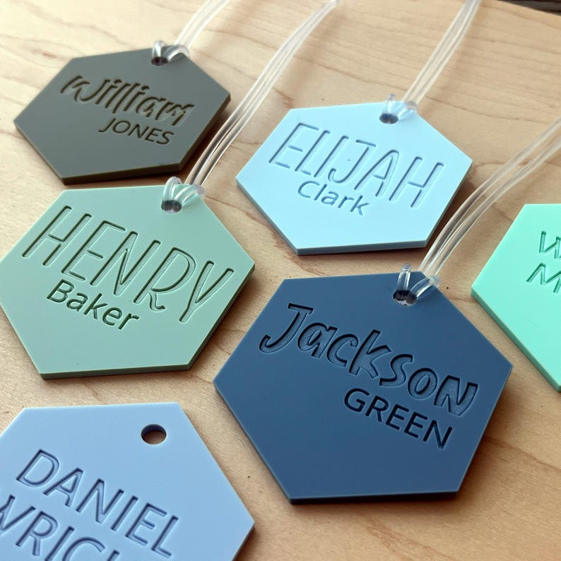 May include: Six hexagonal luggage tags with different names and last names printed on them. The tags are in various colors, including blue, green, and brown. The tags are made of a durable material and have a clear plastic strap for attaching to luggage.