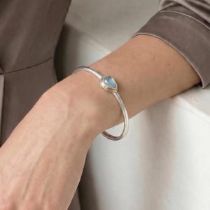 May include: A silver cuff bracelet featuring a single teardrop-shaped light blue gemstone set in a gold-toned bezel. The bracelet has a smooth, polished finish and a simple open-ended design that rests securely around the wrist.