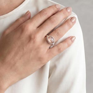 May include: A silver ring featuring a smooth, oval-shaped stone with a translucent, pale pinkish-peach hue. The stone is set in a simple, polished silver bezel band that rests on the ring finger, showcasing a clean and understated design.
