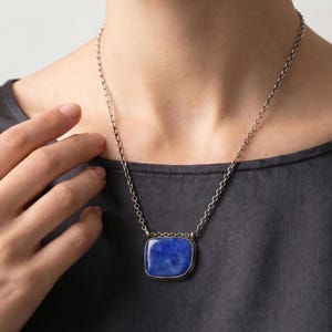 Könnte beinhalten: Eine Halskette mit einem rechteckigen, tiefblauen Steinanhänger in einer silberfarbenen Metallfassung. Der Anhänger hängt an einer mittelschweren silberfarbenen Kette mit abgerundeten Gliedern und liegt im Bereich des Schlüsselbeins.
