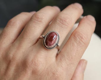 May include: A silver ring featuring an oval-shaped, banded reddish-brown stone set in a twisted rope-style metal bezel. The band of the ring is smooth and polished, and the stone displays concentric circular patterns in shades of deep red and brown.