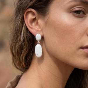 May include: A pair of drop earrings featuring two stacked oval-shaped white stones with subtle grey inclusions, set in a polished silver-toned metal frame. The top stone is smaller and circular, while the bottom stone is larger and elongated.