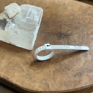 Peut inclure: Un baguier en plastique blanc avec des marques noires pour mesurer les tailles de bague de 3 à 17. Le baguier est sur une surface en bois.