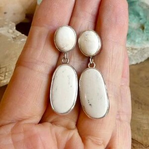 May include: A pair of drop earrings featuring two oval-shaped white stones with subtle grey inclusions, each set in a silver-toned metal frame. The design consists of a smaller oval stone at the top connected to a larger, elongated oval stone below.