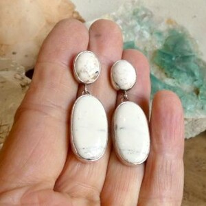 May include: A pair of drop earrings featuring two smooth, oval-shaped white stones with subtle grey veining. Each earring consists of a smaller circular stone at the top connected to a larger, elongated oval stone below, set in a simple metal frame.