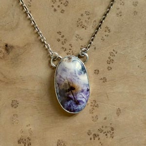 May include: A silver necklace featuring an oval pendant with a marbled pattern of white, purple, and brown hues. The pendant is encased in a silver bezel and hangs from a delicate silver chain. The necklace is displayed on a wooden surface.