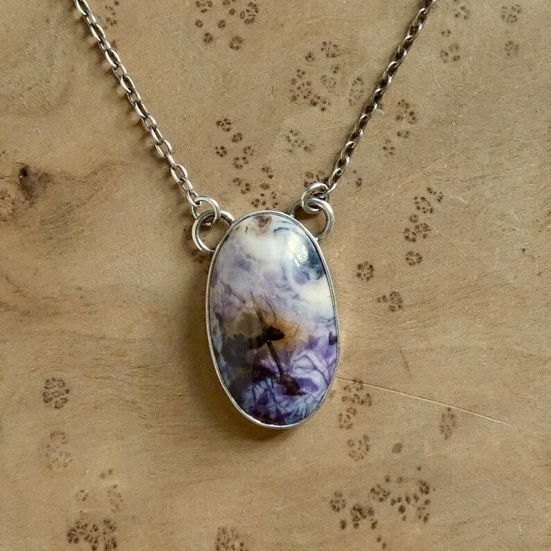 May include: A silver necklace featuring an oval pendant with a marbled pattern of white, purple, and brown hues. The pendant is encased in a silver bezel and hangs from a delicate silver chain. The necklace is displayed on a wooden surface.