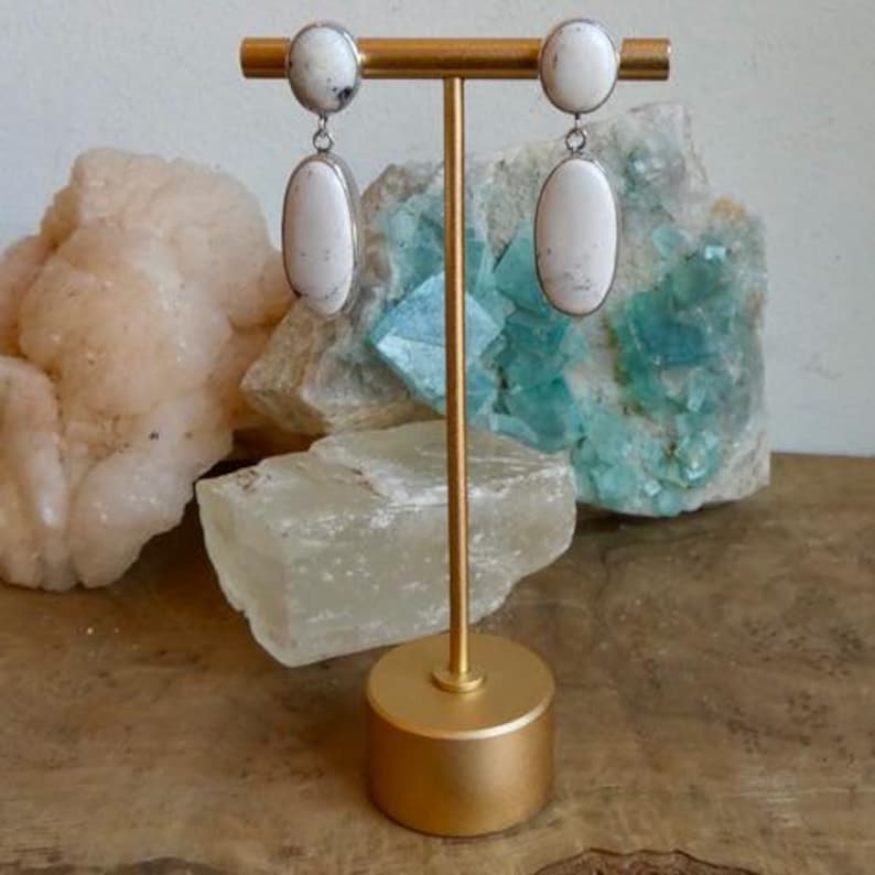 May include: A pair of drop earrings featuring two white oval stones with black inclusions, set in silver-toned metal. Each earring consists of a smaller circular stone at the top and a larger elongated oval stone hanging below, displayed on a gold stand.