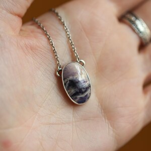 May include: A silver necklace with an oval pendant featuring a polished stone with shades of purple, white, and black. The stone is set in a silver bezel and hangs from a delicate silver chain. The necklace is displayed on a person's open palm.