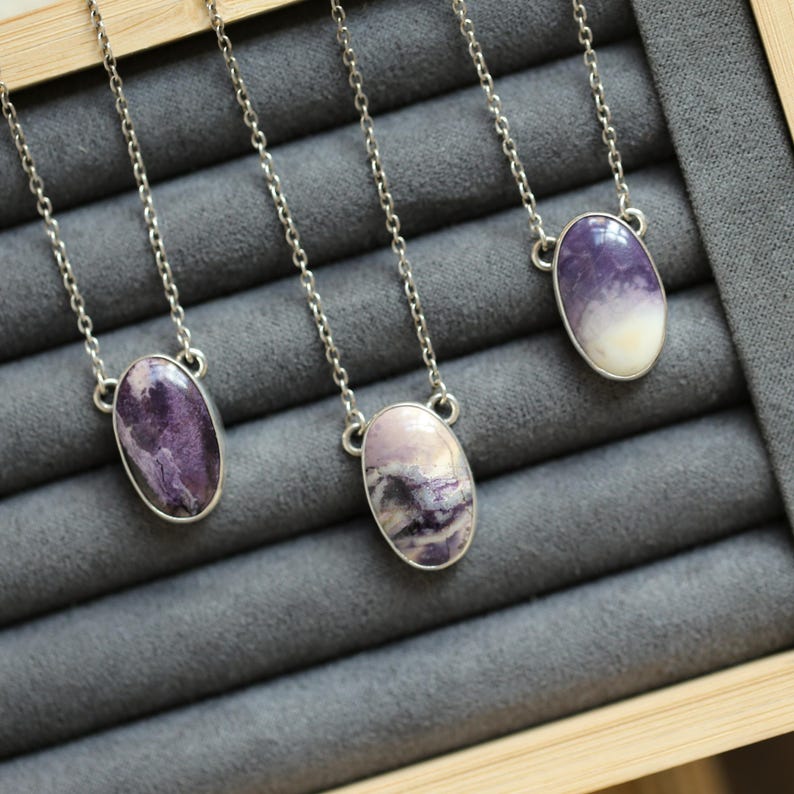 May include: Three silver necklaces with oval-shaped, purple and white stone pendants. The stones have unique patterns and are set in silver bezels. The necklaces are displayed on a gray surface.