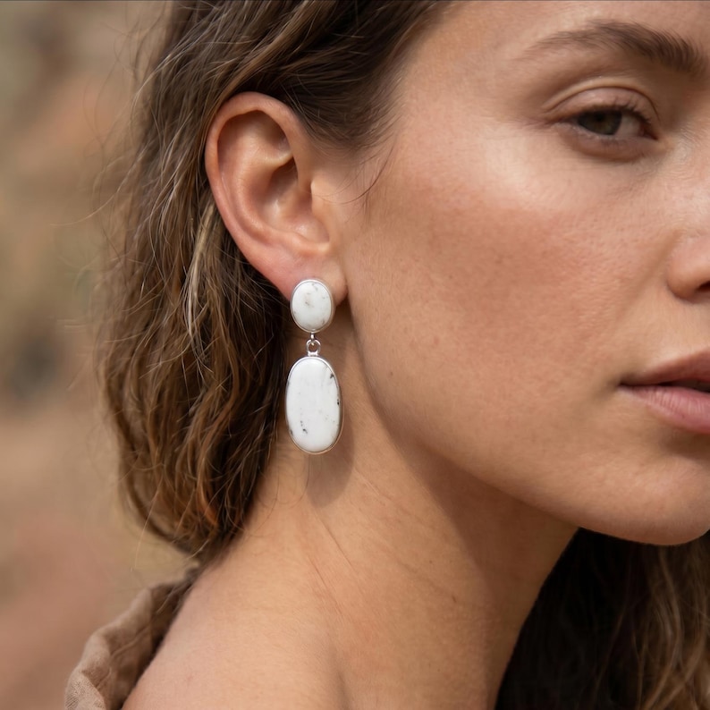 May include: A pair of drop earrings featuring two stacked oval-shaped white stones with subtle grey inclusions, set in a polished silver-toned metal frame. The top stone is smaller and circular, while the bottom stone is larger and elongated.