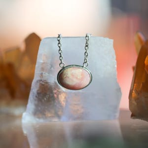K&ouml;nnte beinhalten: Eine Halskette mit einem ovalen, rosa-wei&szlig; gemusterten Steinanh&auml;nger in einer silberfarbenen Metallfassung. Der Anh&auml;nger h&auml;ngt an einer zierlichen silberfarbenen Metallkette mit kleinen, gleichm&auml;&szlig;igen runden Gliedern, die oben an der Fassung befestigt sind.