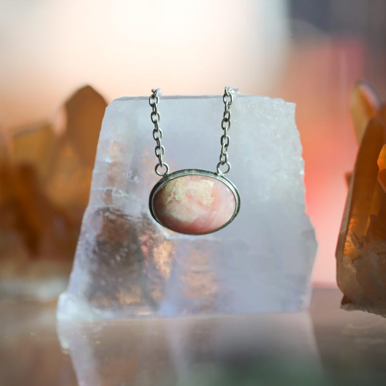 K&ouml;nnte beinhalten: Eine Halskette mit einem ovalen, rosa-wei&szlig; gemusterten Steinanh&auml;nger in einer silberfarbenen Metallfassung. Der Anh&auml;nger h&auml;ngt an einer zierlichen silberfarbenen Metallkette mit kleinen, gleichm&auml;&szlig;igen runden Gliedern, die oben an der Fassung befestigt sind.