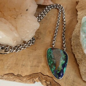 May include: A necklace featuring a silver-toned metal chain and a teardrop-shaped pendant. The pendant contains a polished stone with swirling patterns of deep green, blue, and brown, encased in a smooth, thin silver-toned metal border.