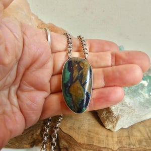 May include: A pendant necklace featuring an oval-shaped azurite-malachite gemstone with swirling patterns of deep blue, green, and brown. The stone is set in a smooth silver bezel and hangs from a thick, round silver link chain.